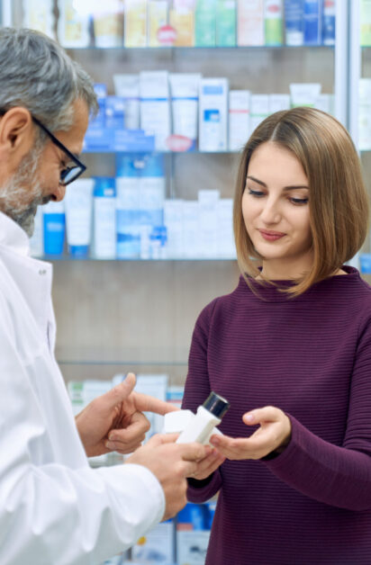 Pharmacist offering pretty female customer medical products in drugstore. Young woman holding white bottles, choosing product. Side view of mature chemist wearing glasses and white lab coat.